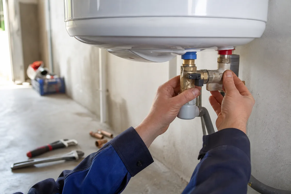 Chauffe-eau électrique dans le garage d’un pavillon, mains d’un plombier vérifiant le groupe de sécurité