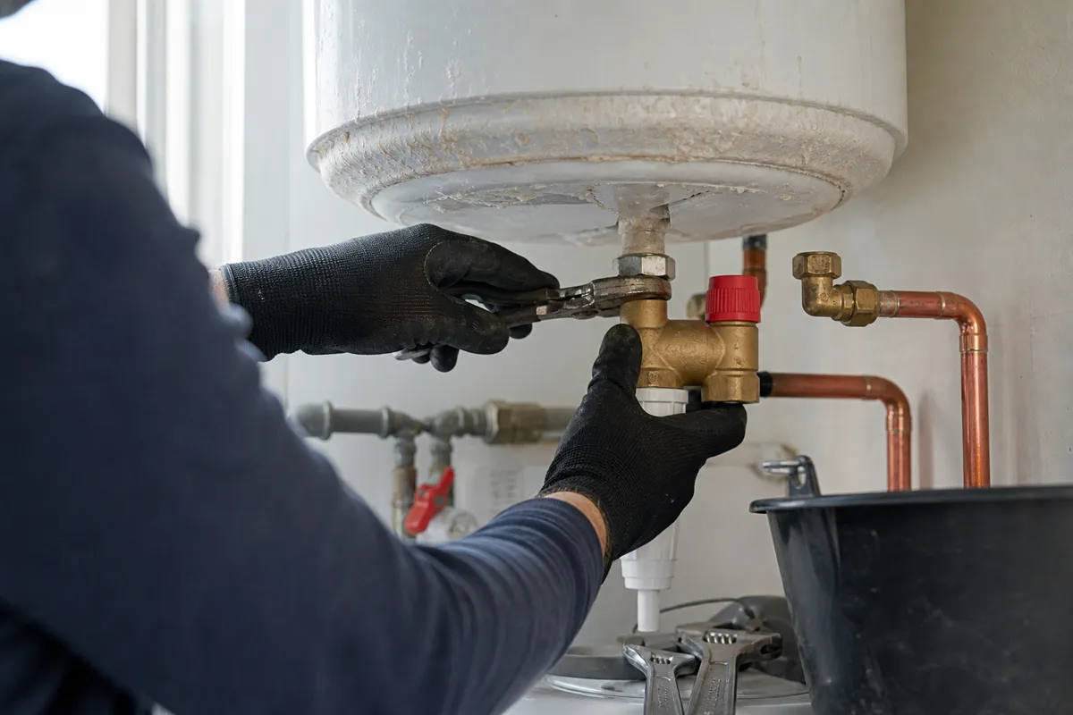 Plombier remplaçant un groupe de sécurité sous un cumulus électrique entartré, mains gantées et outillage propre